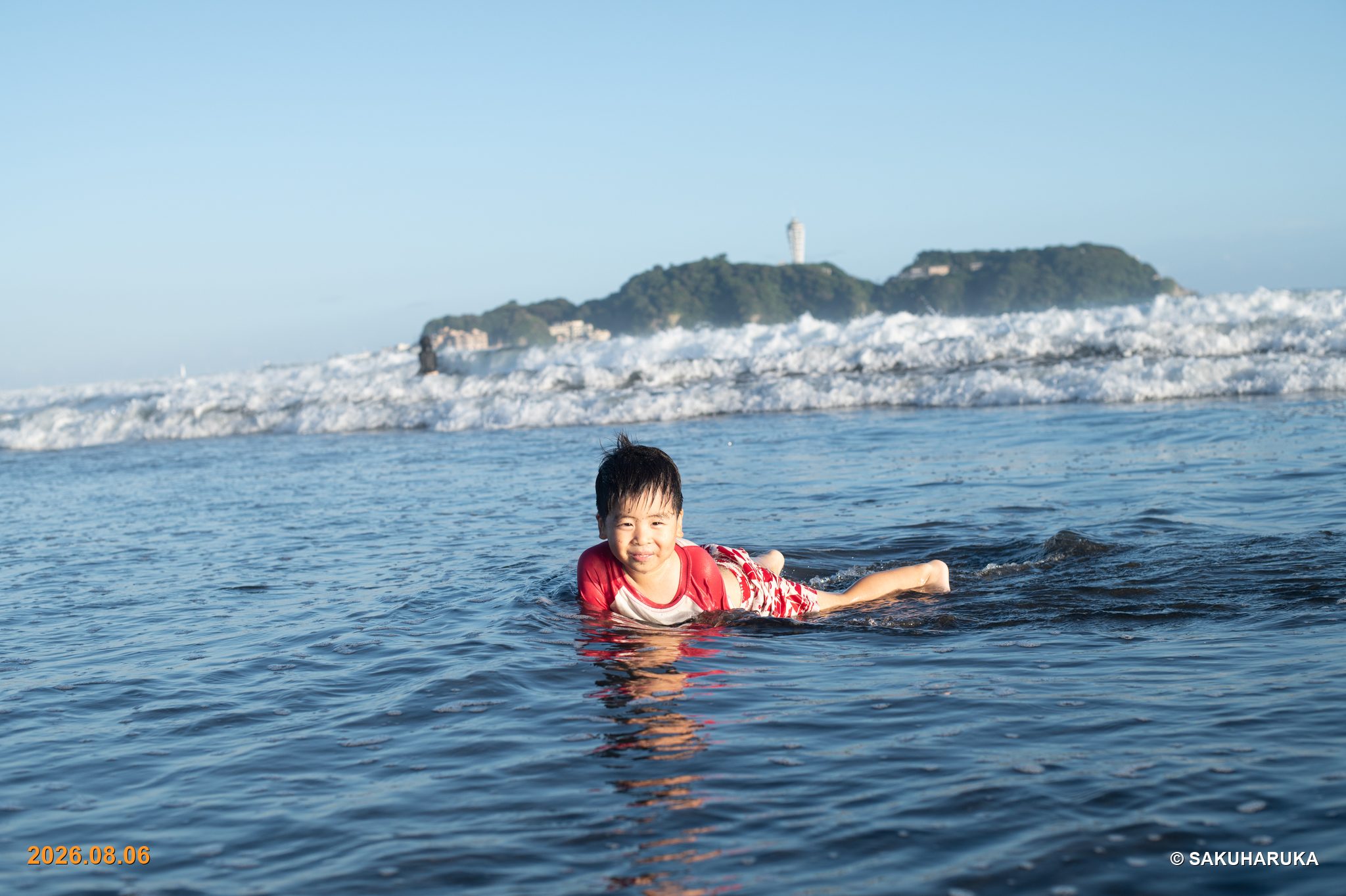 江の島を背に、浅い波に腹ばいになって笑う男の子